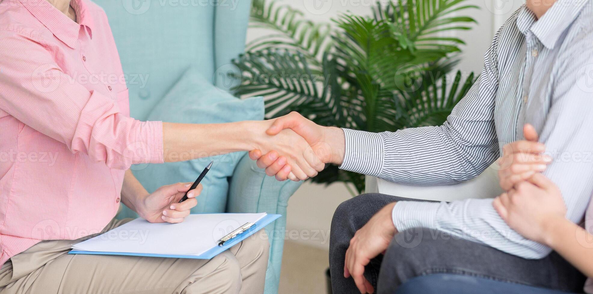 A therapist and a client shake hands during a consultation in a comfortable, warmly-lit office. The therapist holds a clipboard and pen, cropped, panorama photo