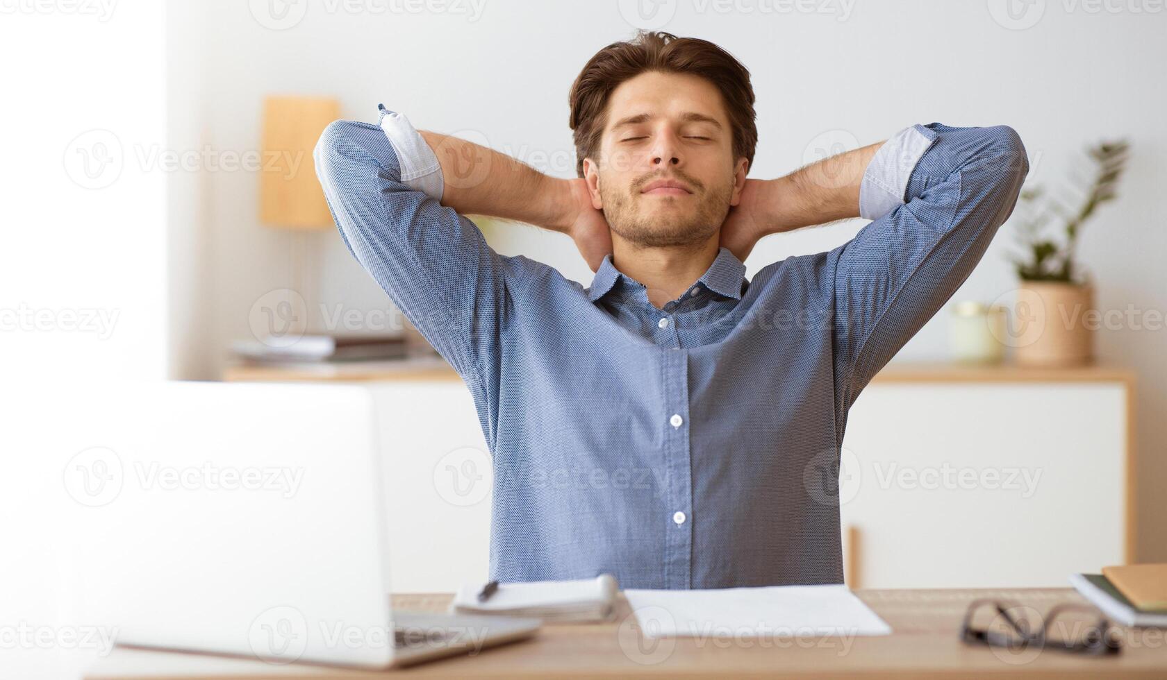A man sits at his desk with a laptop, eyes closed and hands behind his head, taking a moment to relax. photo