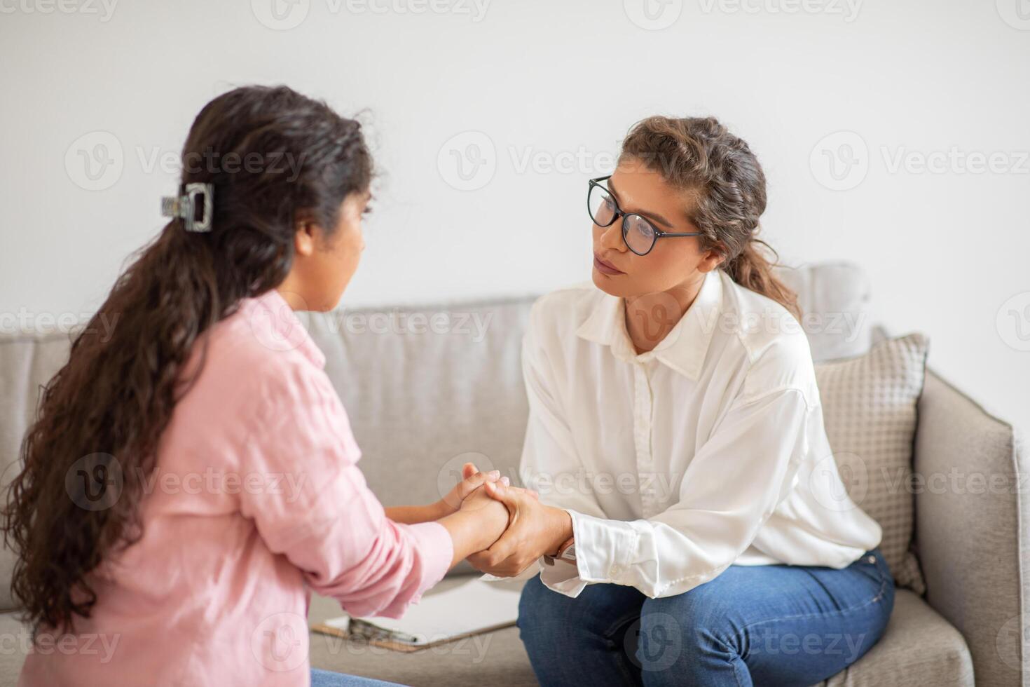 A woman is offering support and comfort to another woman who is sitting on a couch. She is holding the woman hand and looking at her with concern. photo
