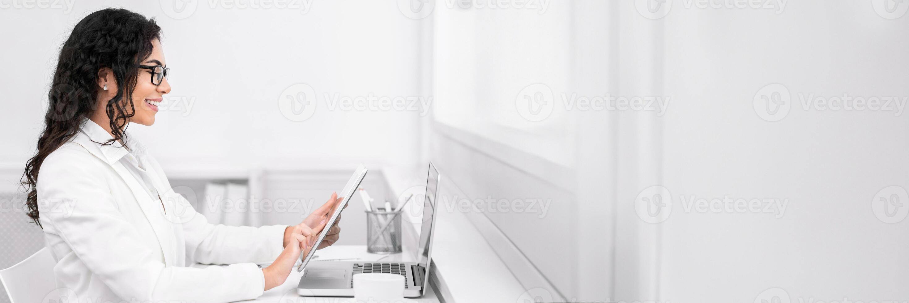 Break Time. Side view of mexican woman reading story on e-book at work, holding tablet in front of laptop, copy space photo