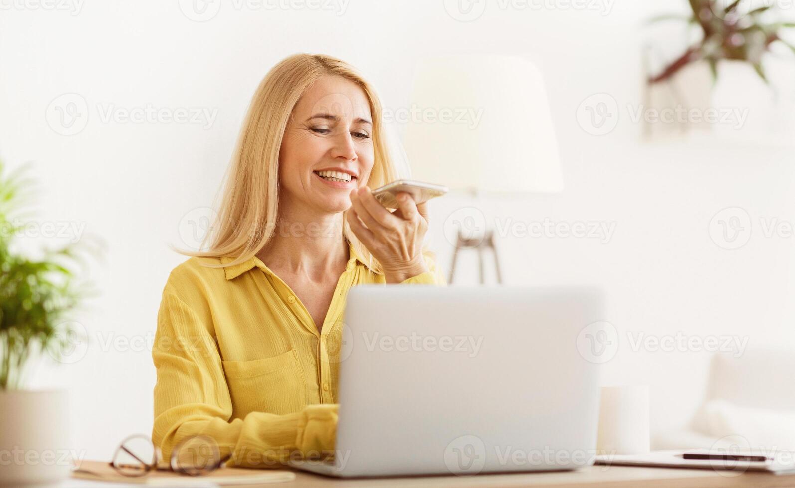 A woman with blonde hair sits at a desk using a voice assistant while working on a laptop. photo