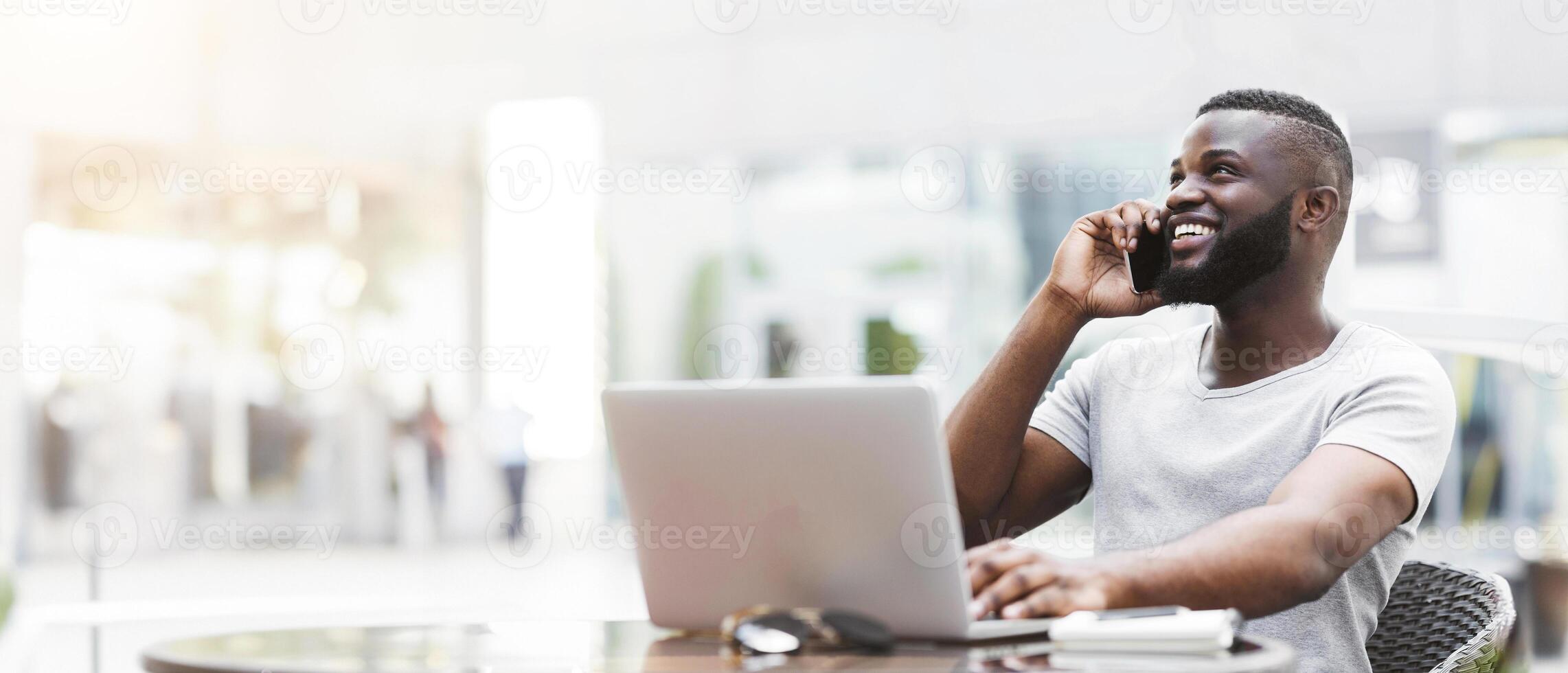A man sits at a table with a laptop open, smiling and talking on his phone. photo
