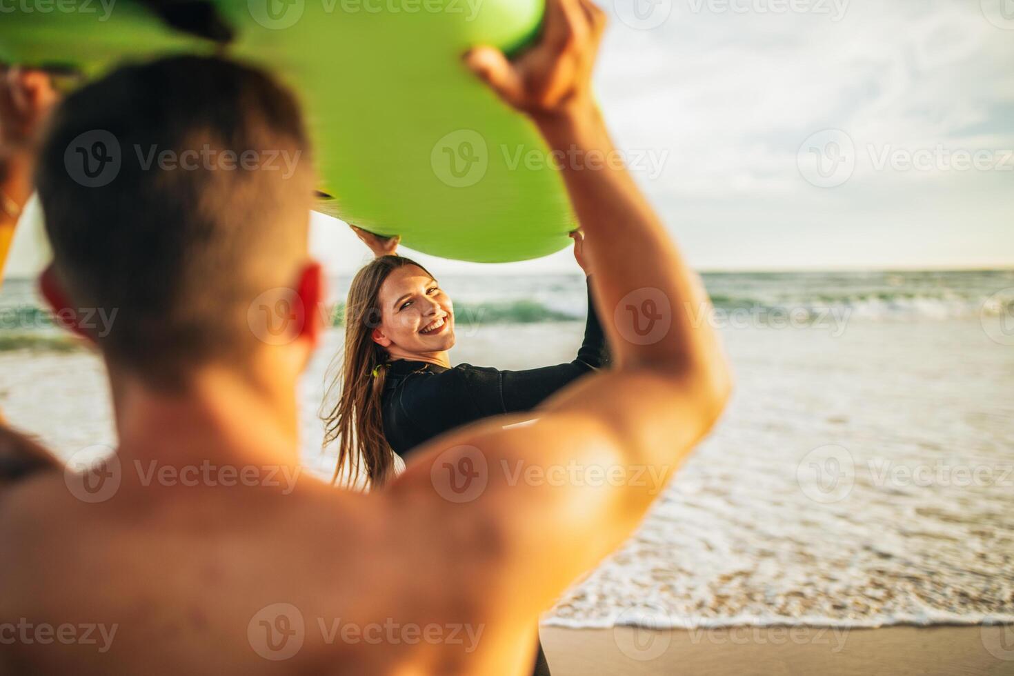A couple is standing on a sandy beach with a green surfboard held above their heads. The man is facing away from the camera and the woman is looking back at the camera, smiling. photo