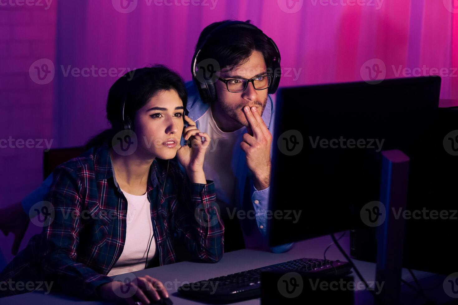 A couple wearing headphones sits at a computer desk at night, intently watching a monitor. They are both engaged in the activity photo