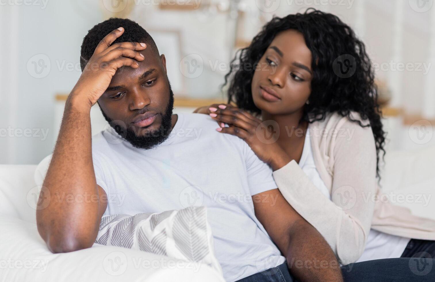 A woman sits beside a man on a couch, her hand resting on his shoulder as he sits with his head in his hand, looking down. photo
