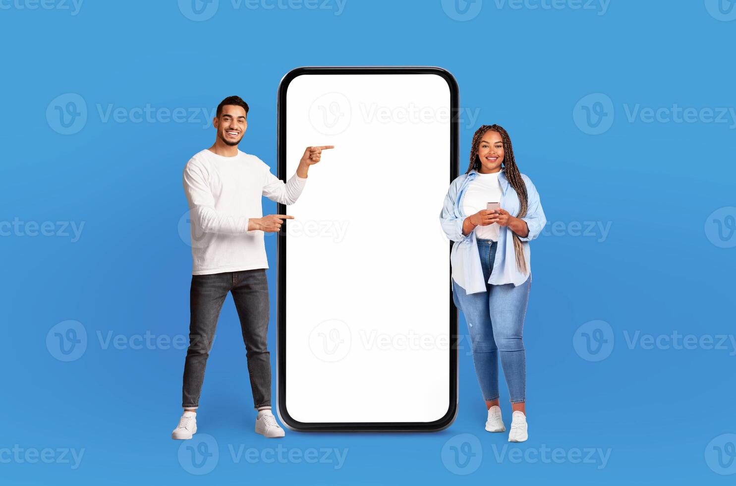 Multiethnic man and woman are standing side by side next to large phone, looking at the screen and appearing to have conversation. The phone is significantly bigger than them, emphasizing its size. photo