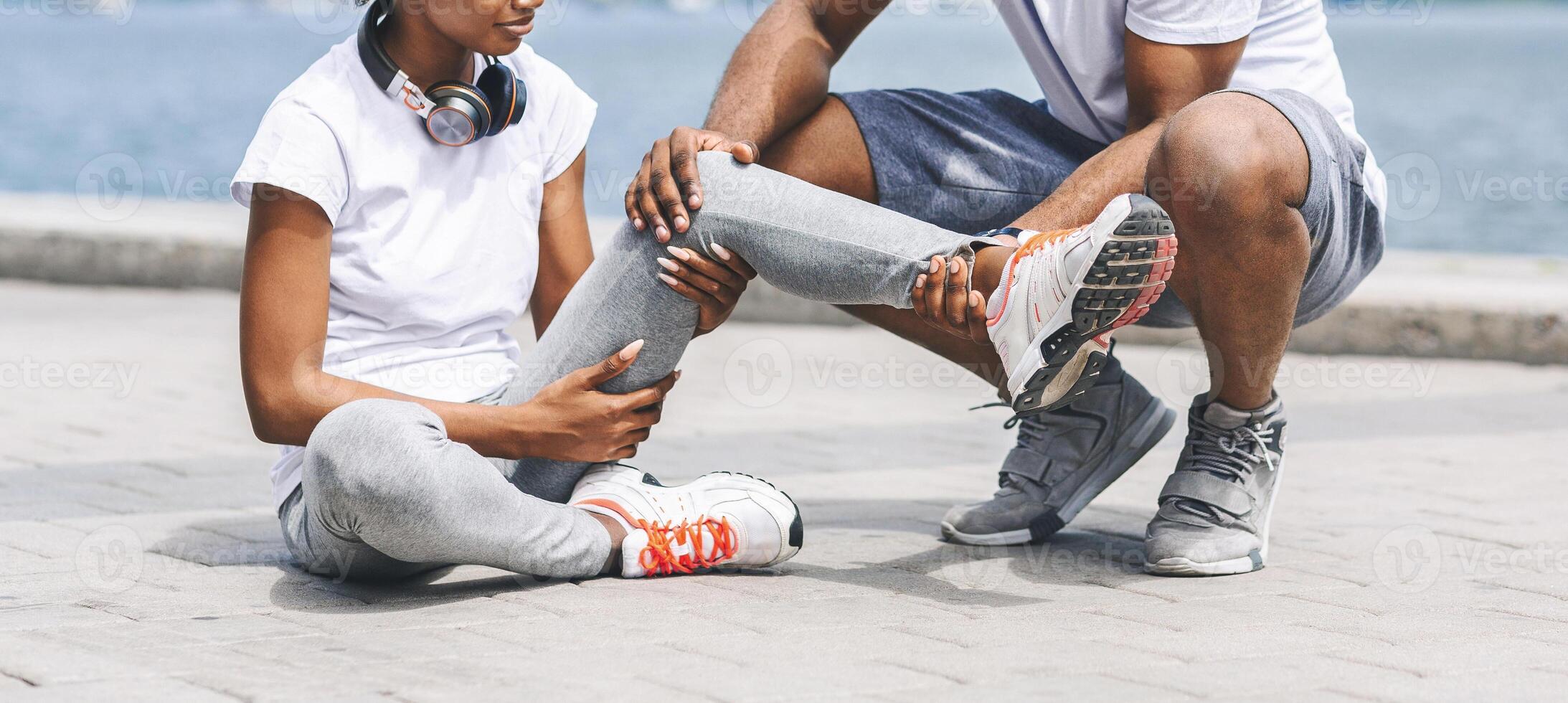 Jogging Injury Concept. Unrecognizable Afro Man Helping Woman With Injured Knee Sitting Near River. Cropped, Panorama photo