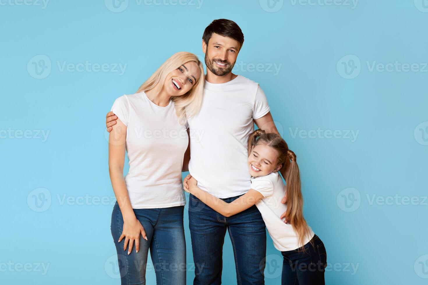A happy family of three poses for a portrait against a light blue background. The parents are both smiling and looking at the camera, while their young daughter hugs her father from behind. photo