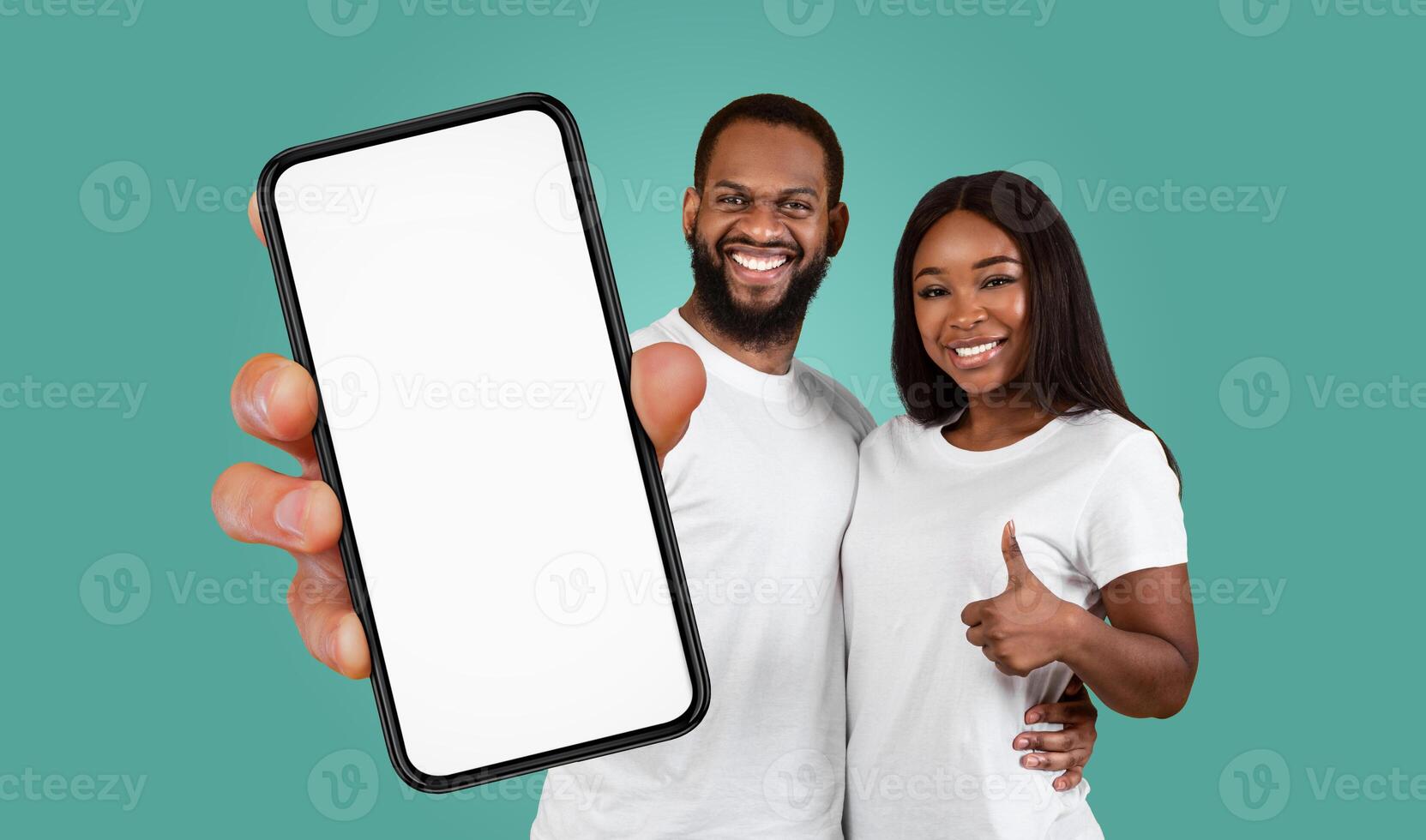 A young Black couple is standing in front of a teal background. The man is holding a smartphone with a blank white screen in front of him, and the woman is giving a thumbs up, smiling. photo