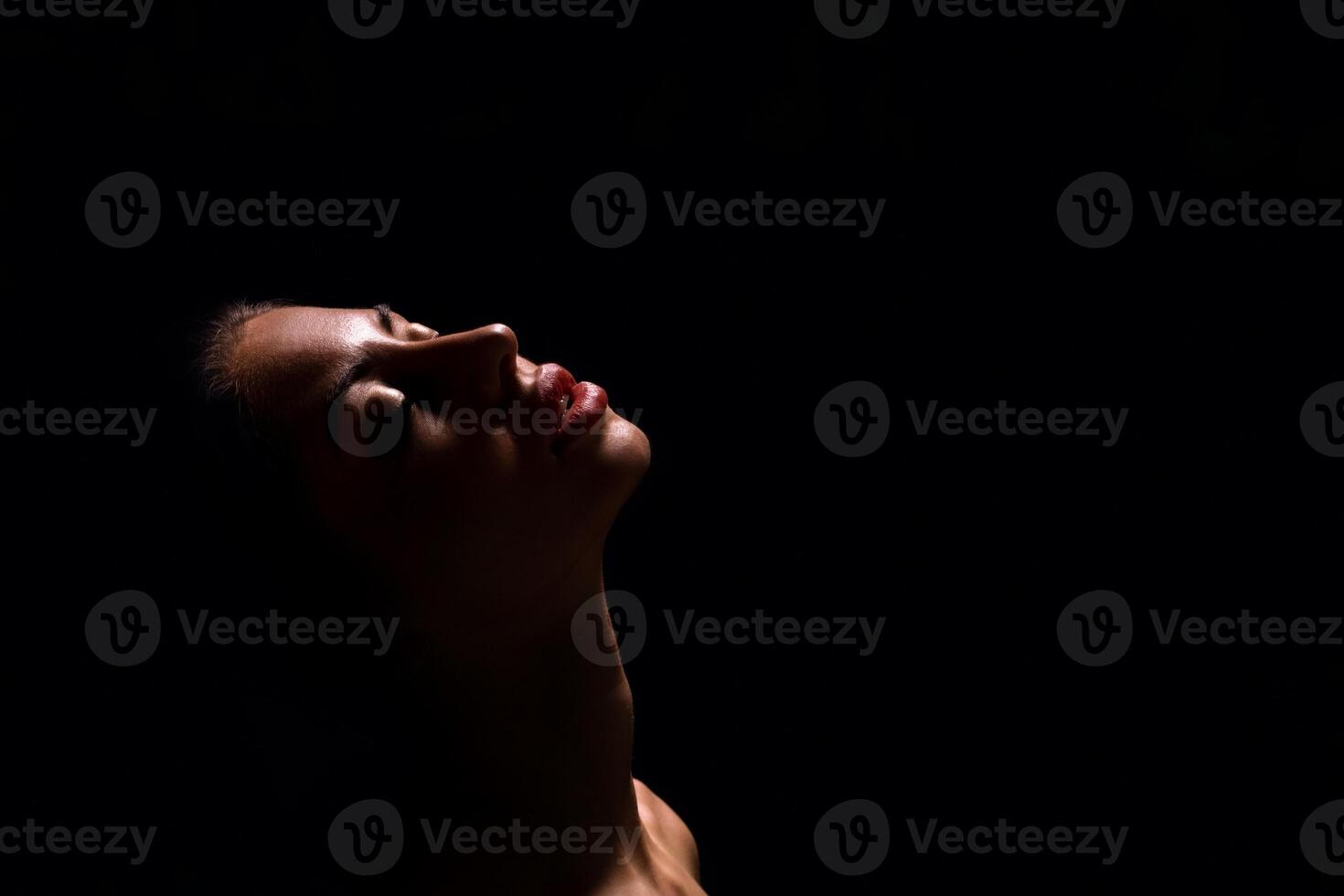 A close-up shot of a womans face, illuminated by a single light source, with her eyes closed and lips slightly parted. The background is completely black, copy space photo