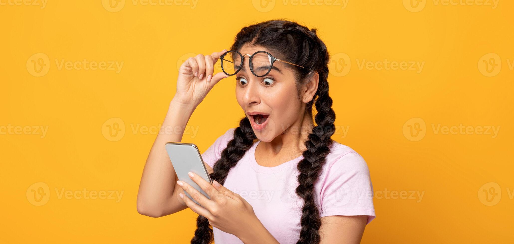 Wow, Great News. Portrait Of Excited Surprised Indian Woman Holding And Using Smartphone, Browsing Social Media. Emotional Young Lady Having Success, Taking Off Glasses, Yellow Studio Background photo
