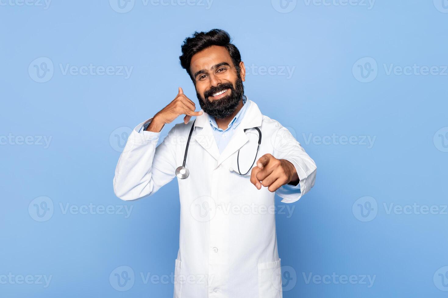 Indian doctor with a beard is smiling and making a call me gesture with his left hand while pointing his right index finger forward. He is standing in front of a blue background. photo