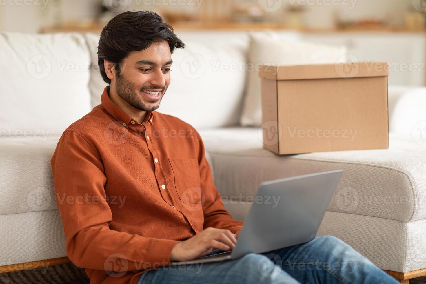 Arab man is sitting on a couch while using a laptop computer. He is focused on the screen, typing and scrolling. The room is cozy with a coffee table and a lamp in the background. photo