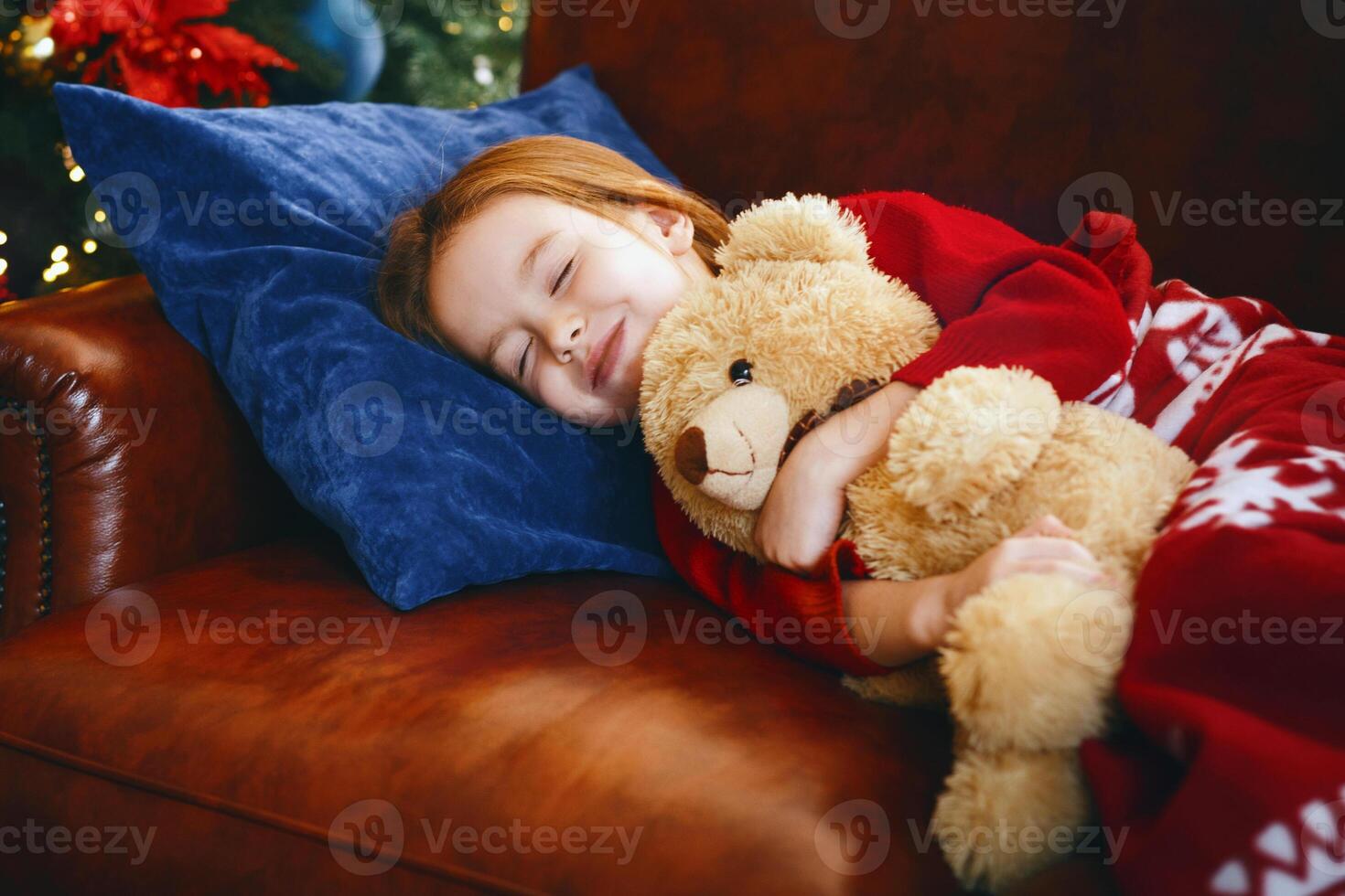 Cute little girl sleeping next to illuminated Christmas tree, dreaming about christmas miracles, hugging teddy bear, free space photo