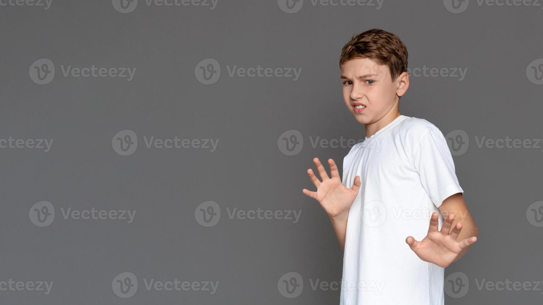 A teenager in a white shirt stands with a defensive posture, expressing caution or fear, against a grey, isolated background. His expression suggests hesitation or concern photo