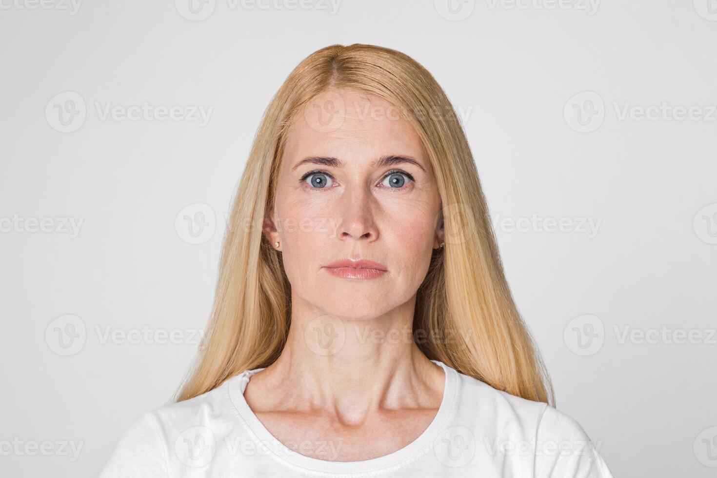 This image shows a woman with blonde hair looking directly at the camera with a neutral expression. She is wearing a white t-shirt and has a light complexion. The background is a plain white wall. photo