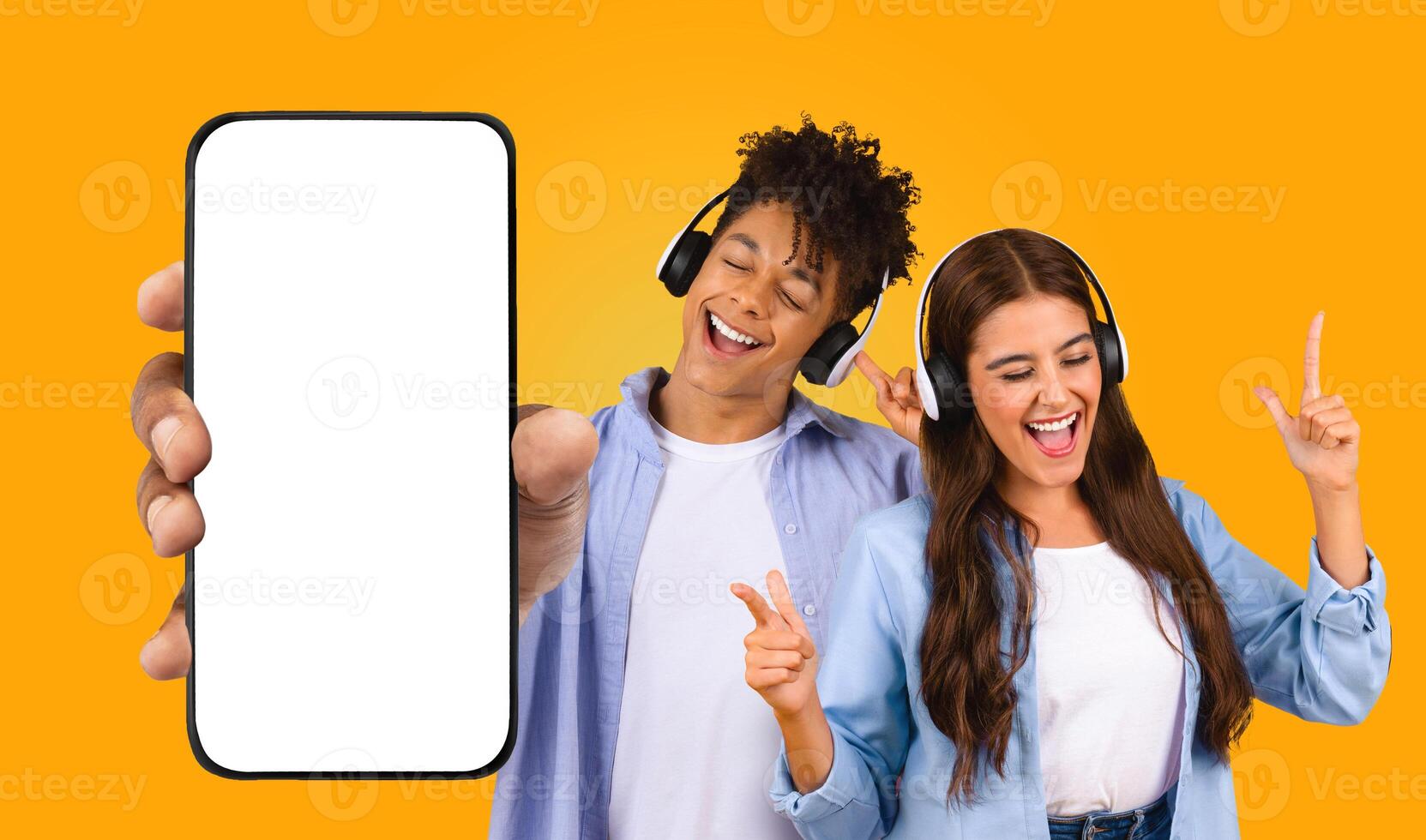 Black man holds a smartphone with a blank screen in front of him while Middle Eastern woman dance and sing along to music on their headphones. They are all happy and enjoying the music. photo