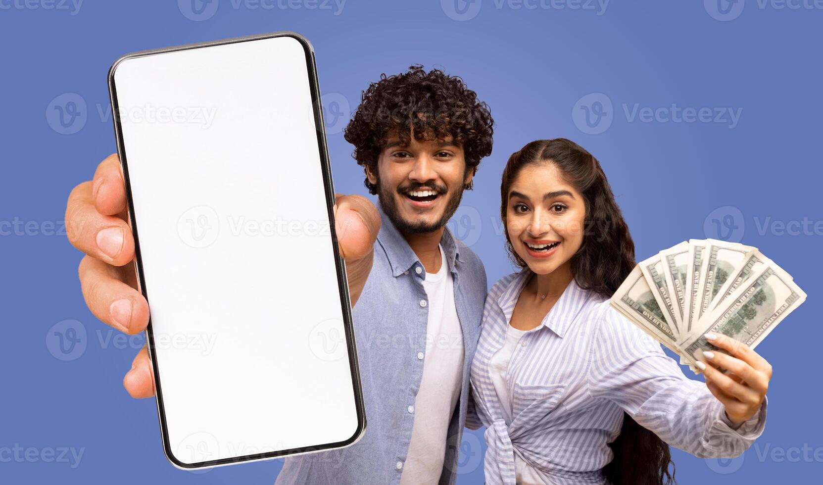 A young Indian couple is holding a smartphone with a blank screen and a fan of money, both facing the camera and smiling. photo