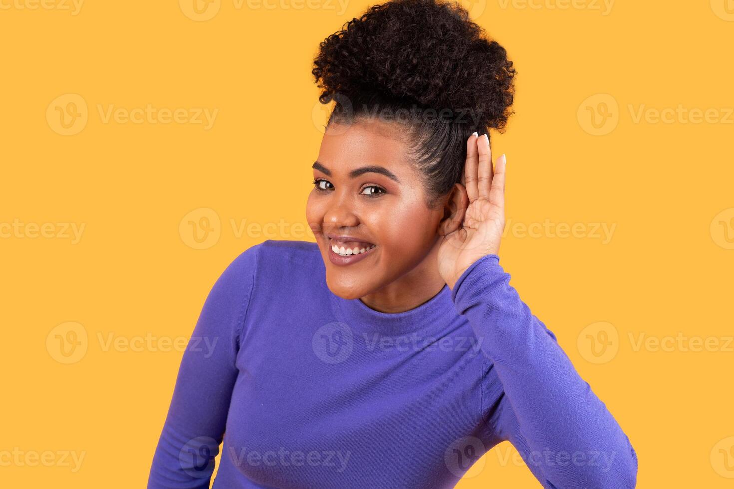 A cheerful young woman raises her hand to her ear, displaying a friendly smile. She is positioned against a vibrant yellow background, creating a lively and engaging atmosphere. photo