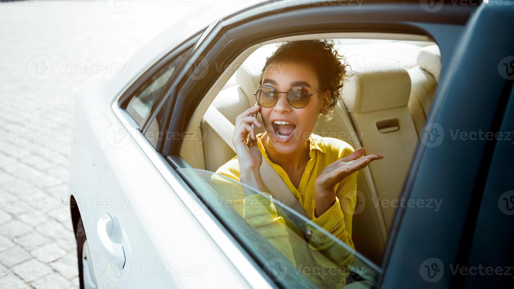 A woman with curly hair and sunglasses is sitting in the back seat of a white car. She is on a phone call and looks excited, with her mouth open in surprise. photo