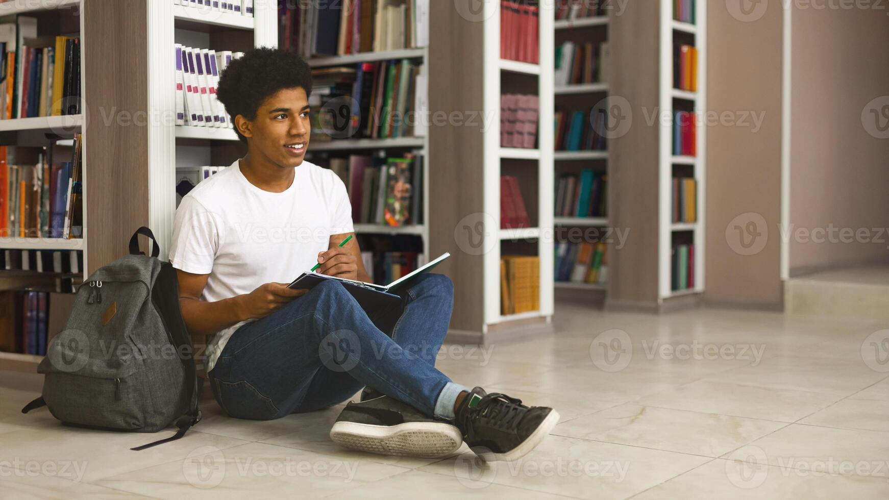 Creative african american student sitting and making notes on the floor at library, free space photo