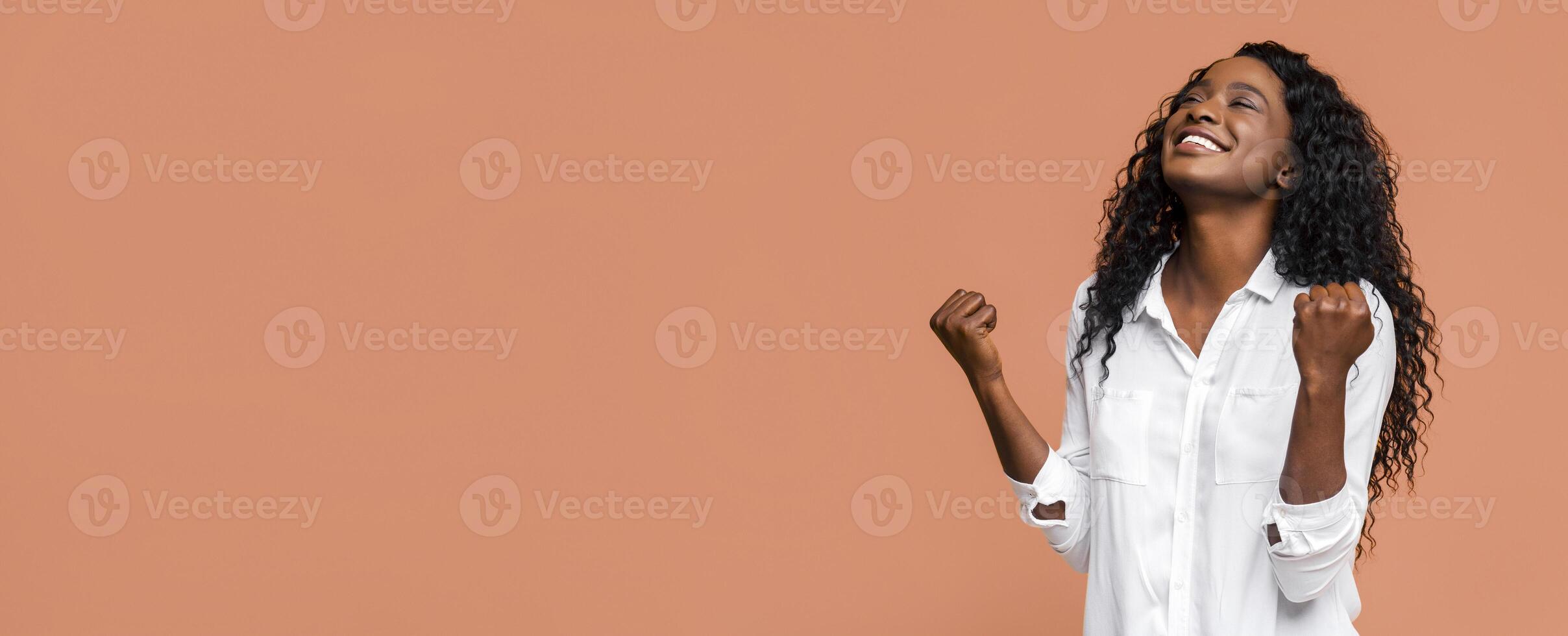 A young Black woman is celebrating her success. She is wearing a white shirt and has her arms raised in the air with her fists clenched. She is smiling and looking up towards the sky. photo