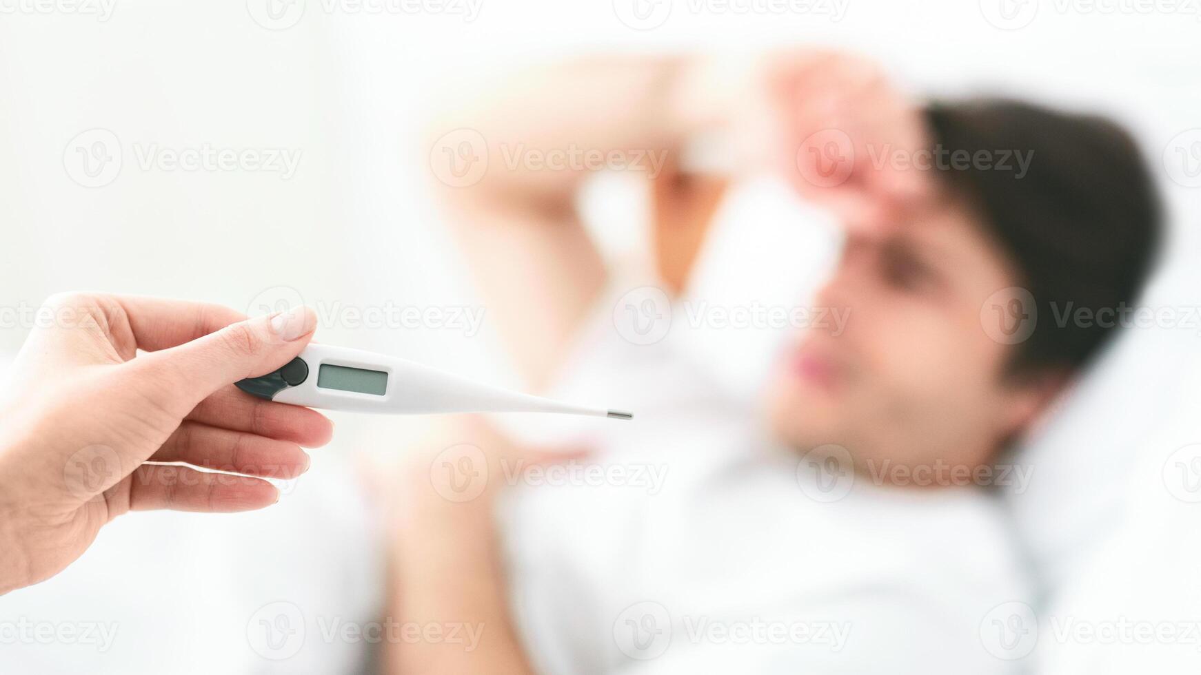 A hand holds a digital thermometer while pointing it at a man lying in bed. The man is out of focus and appears to be sick. photo