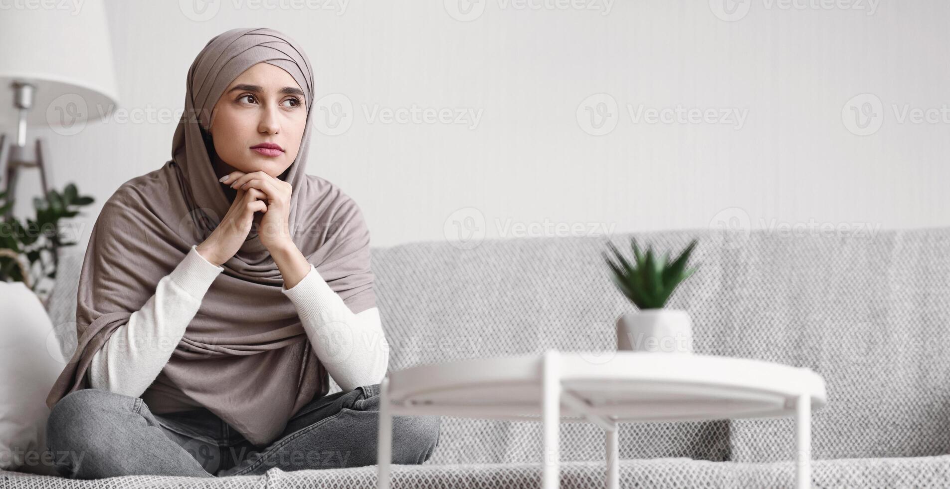 Portrait of thoughtful arabic woman in hijab sitting on couch at home, resting head on hands and looking away with sad face expression, copy space photo
