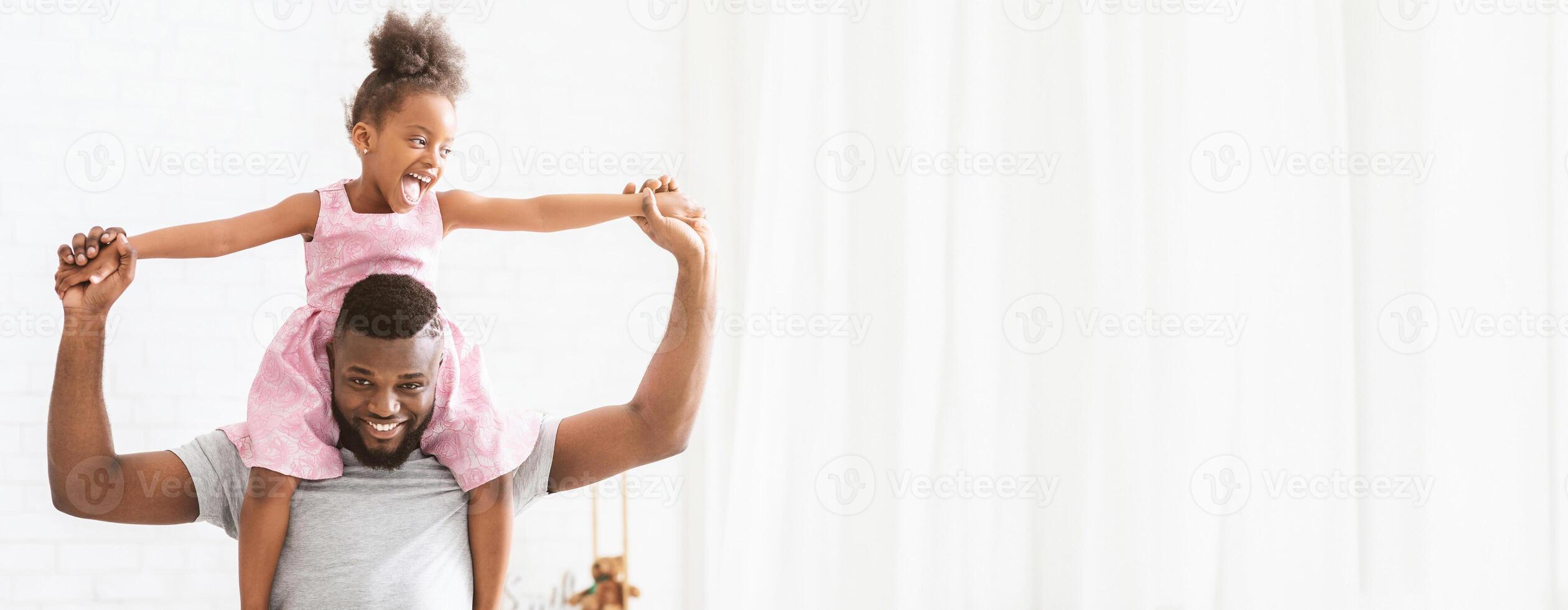 African American father carries his young daughter on his shoulders, both radiating happiness. They are indoors, enjoying playful moments together in a bright and cheerful setting, copy space photo
