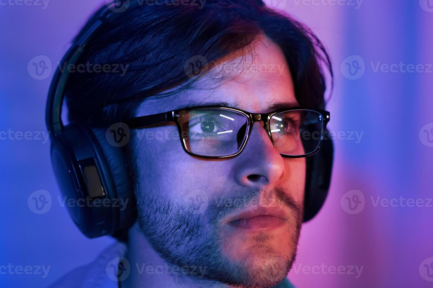 A close-up portrait of a young man wearing glasses and headphones. He is illuminated by blue and pink lighting, and his expression is serious and focused photo