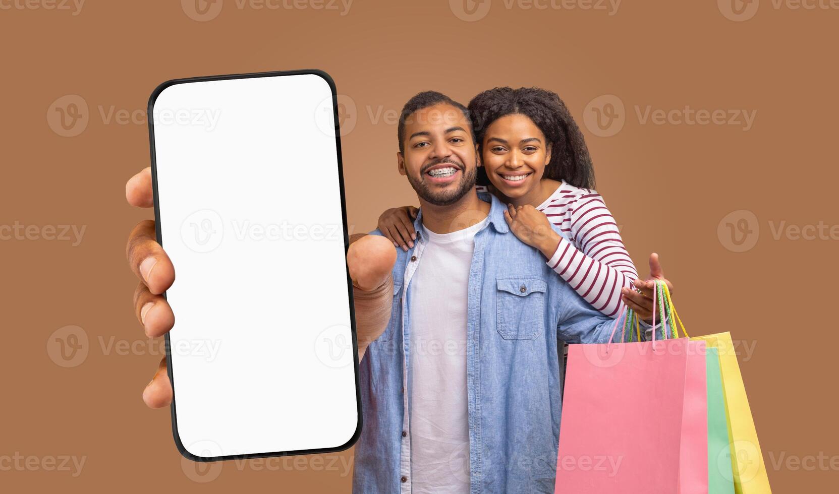 A smiling black couple with shopping bags stands in front of a brown background. The man is holding a large smartphone with a blank screen, pointing to the device with his index finger photo