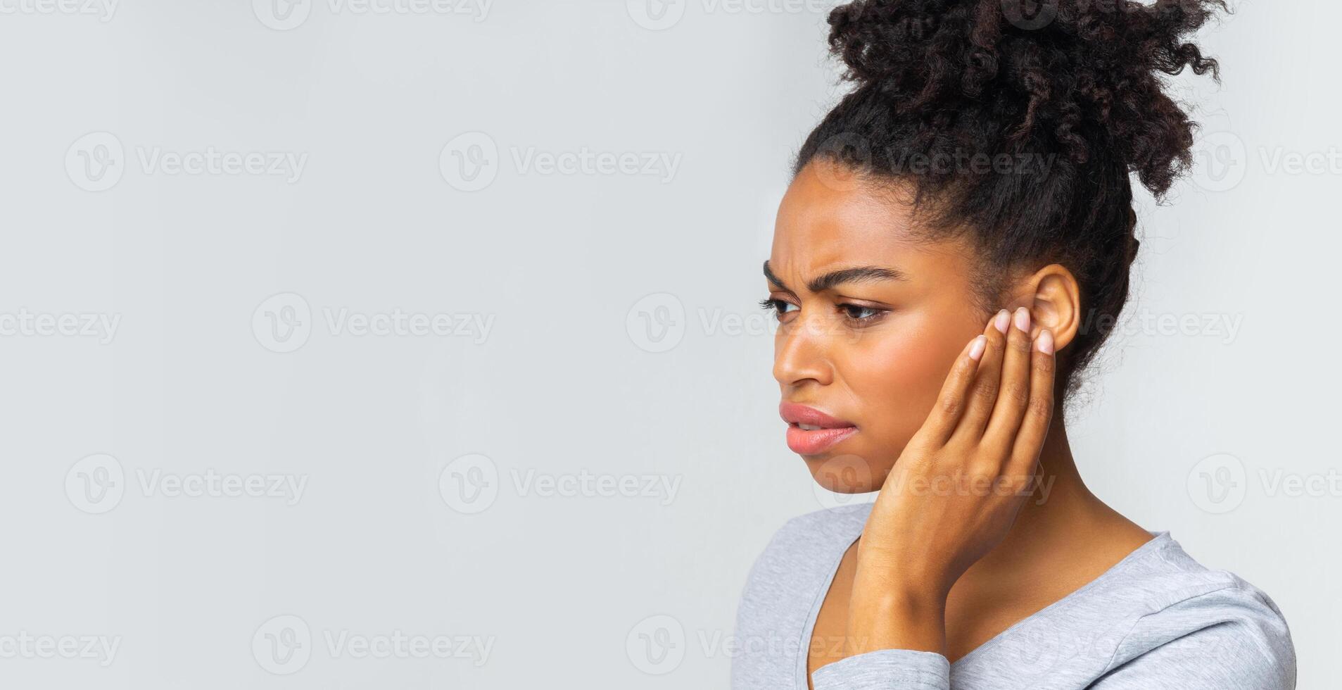 African girl suffering from otitis, rubbing her inflamated ear over grey background, ear diseases concept photo