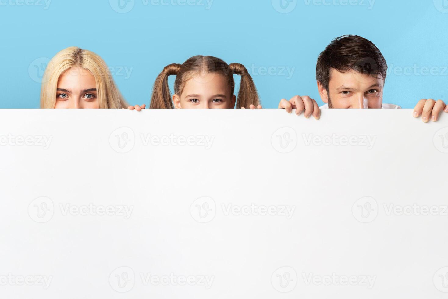 A family, consisting of a mother, father, and daughter, are peeking over a large white sign. They are smiling and looking at the camera. The background is a light blue color. photo