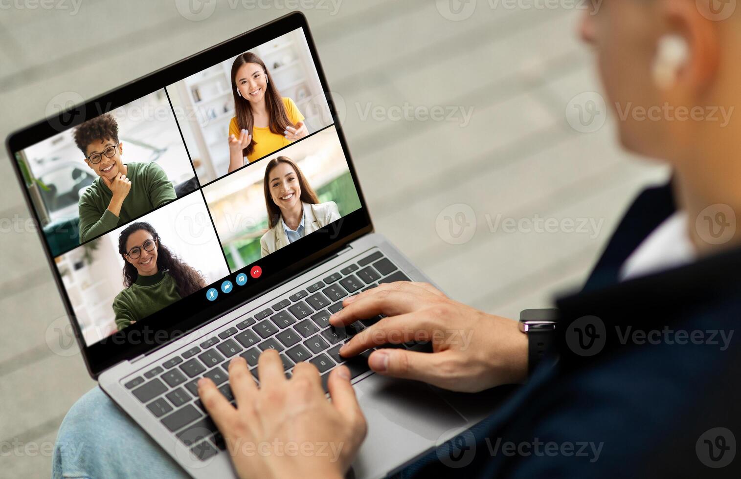 A man is sitting in a chair, using a laptop computer to participate in a conference call. He is typing on the keyboard and looking at the screen, which displays four other people. photo