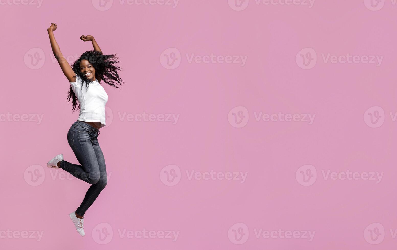 A joyful young woman with dark curly hair is mid-air, jumping with her arms raised in the air. She is wearing a white T-shirt and dark blue jeans. The background is a solid, light pink. photo