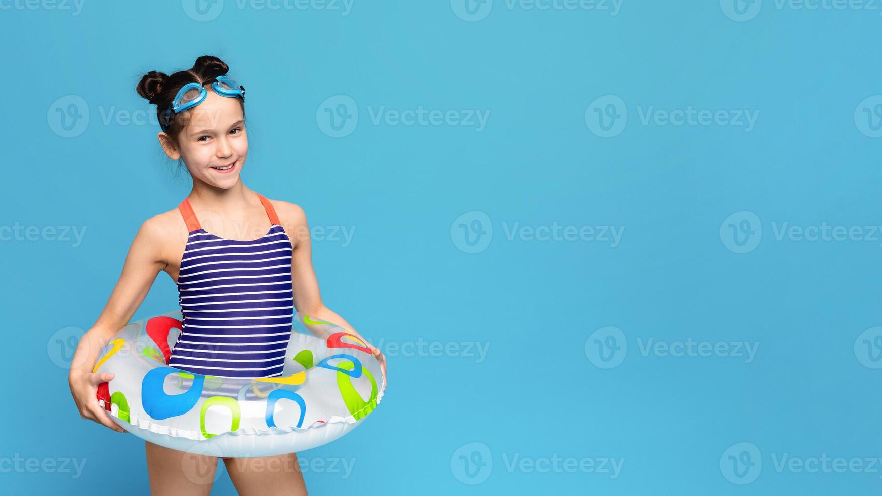 A young girl wearing a colorful bathing suit stands holding an inflatable pool float. The girls hair is tied up in a ponytail as she grips the float with a smile on her face, copy space photo