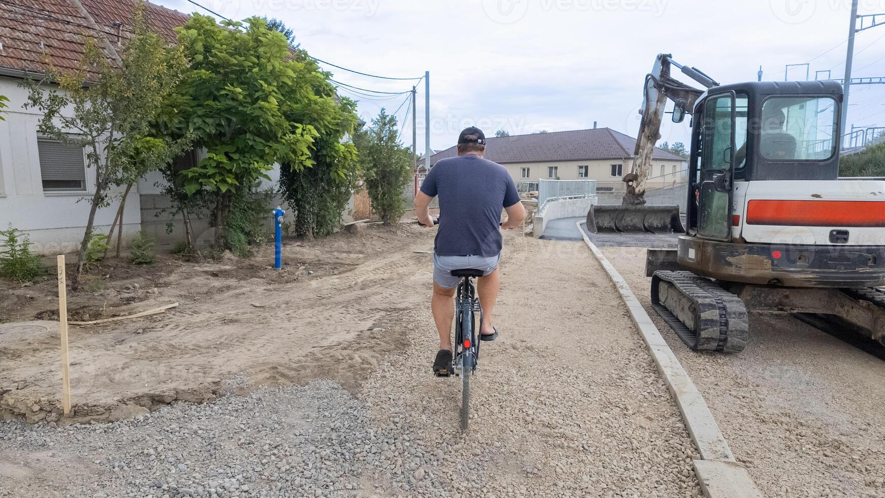 A Caucasian man rides a bicycle on a gravel road next to construction equipment, symbolizing urban development challenges photo