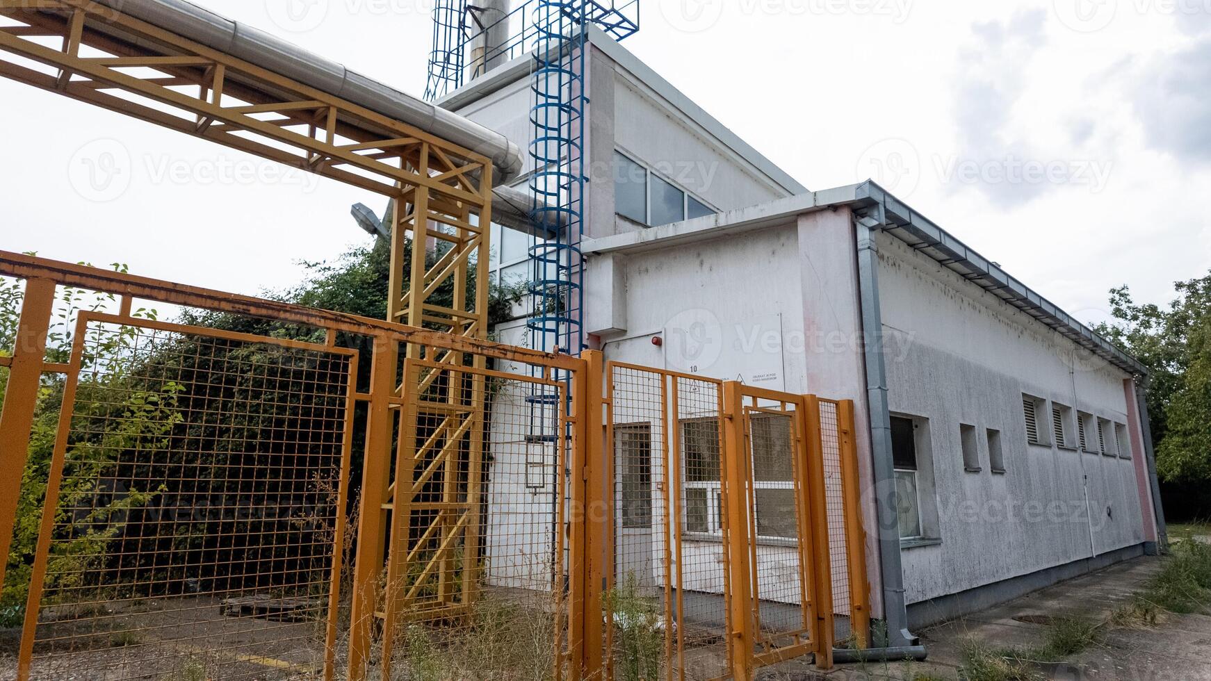Abandoned industrial building with rusting metal fence and overgrown weeds, symbolizing urban decay and economic decline photo