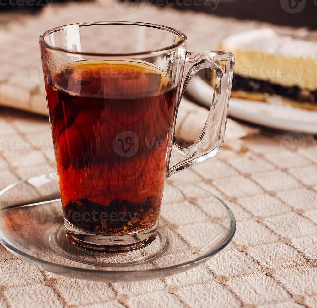 Steaming glass of black tea resting on tablecloth with cake in background photo