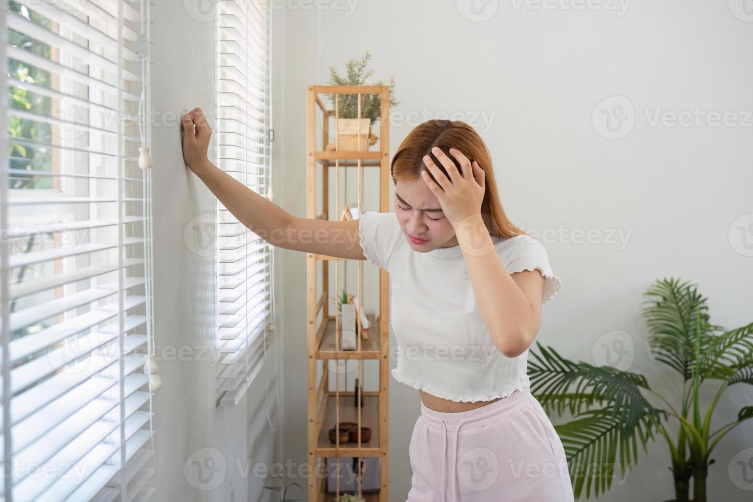 Young Woman Experiencing a Headache at Home, Leaning Against Window with Hand on Forehead, in a Bright and Modern Living Space photo