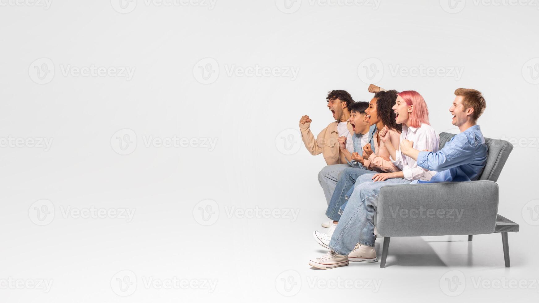 A group of five friends sit on a gray couch, excitedly watching a television screen that is off-camera. The friends are all dressed casually and have a variety of hairstyles, copy space photo