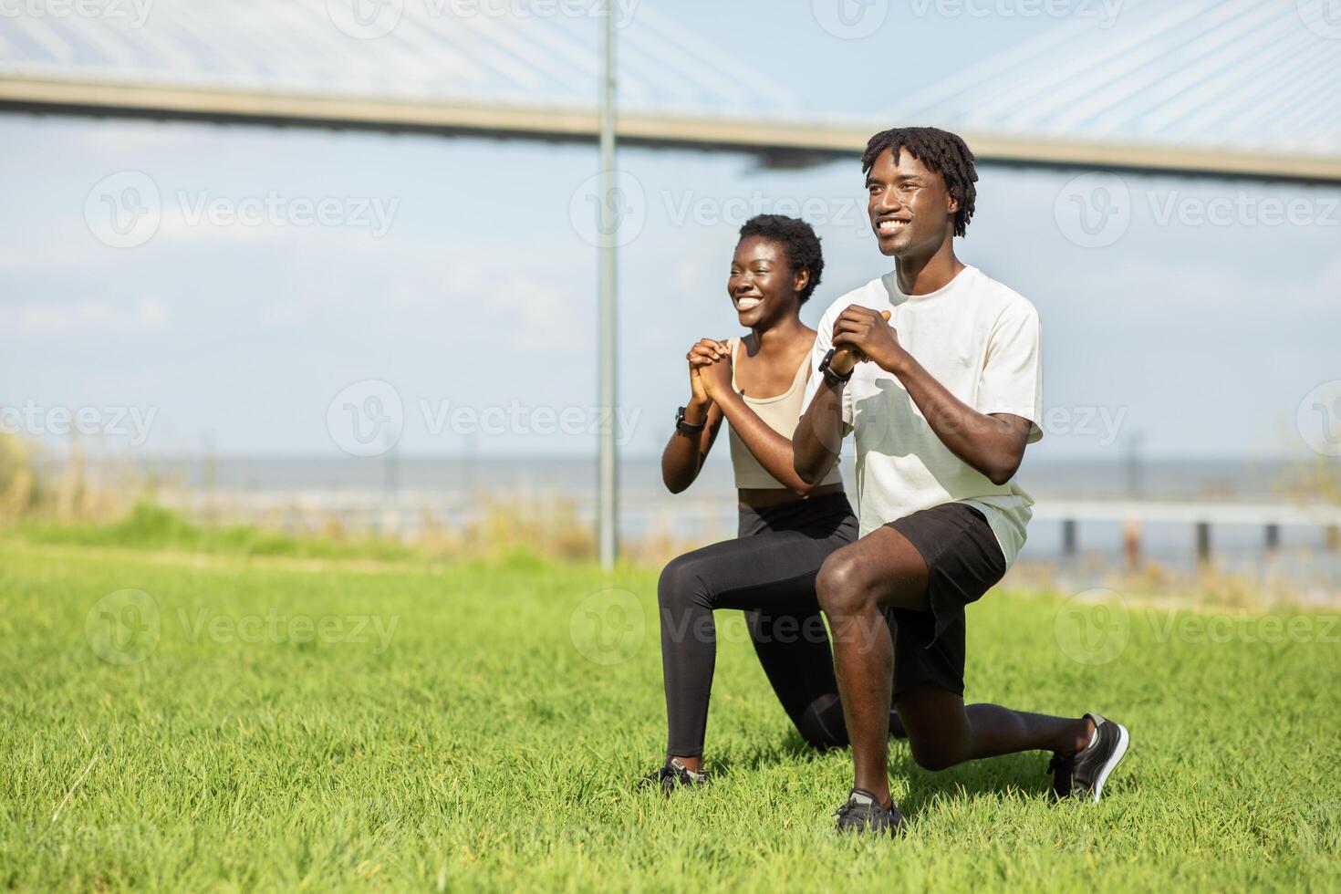 African American couple performs lunges in a grassy area with a bridge and water in the background. They enjoy a sunny day while staying fit and active together. photo