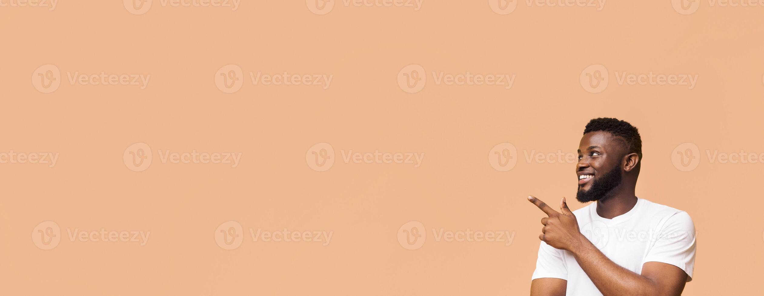 An African American man with a beard smiles and points to the left side of the frame. He is wearing a white t-shirt and standing against a tan background, copy space photo