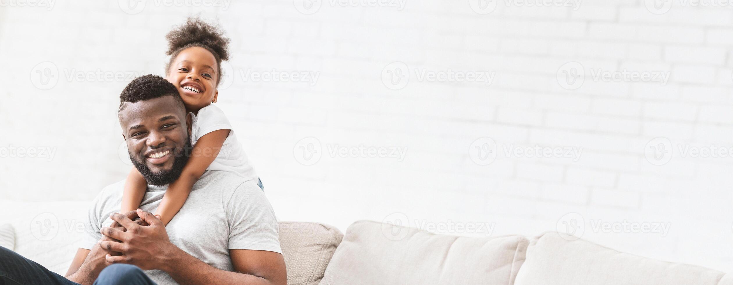 A joyful black father interacts with his young daughter, who playfully hugs him from behind while they sit on a comfortable sofa. The warm atmosphere highlights their close bond, copy space photo