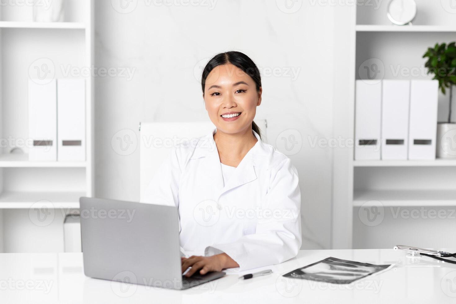 A healthcare professional Asian lady is sitting at a desk, engaging with a laptop while maintaining a cheerful demeanor. Medical files are organized nearby, reflecting a busy yet orderly environment. photo