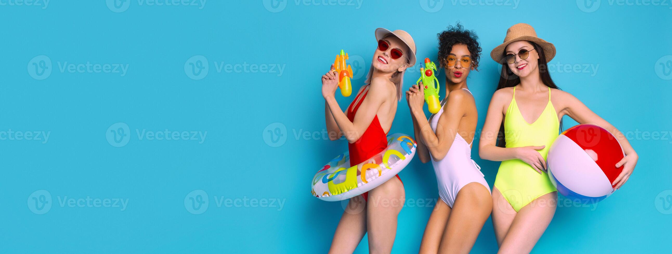 Three women are posing in swimsuits with water guns and a beach ball on a bright blue background. They appear to be having fun and enjoying the summer, copy space photo
