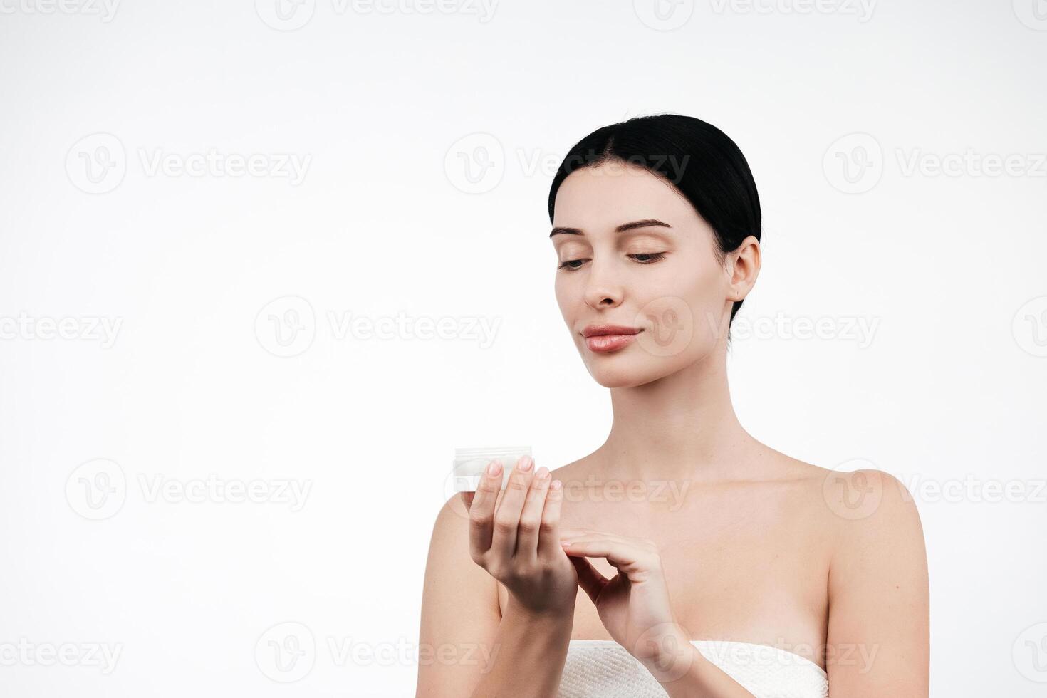 Beauty portrait of brunette woman in white towel, applies cream to the face. Skincare cleansing eco organic cosmetic spa relax concept. Free copy space for text, banner. Cosmetics advertising photo