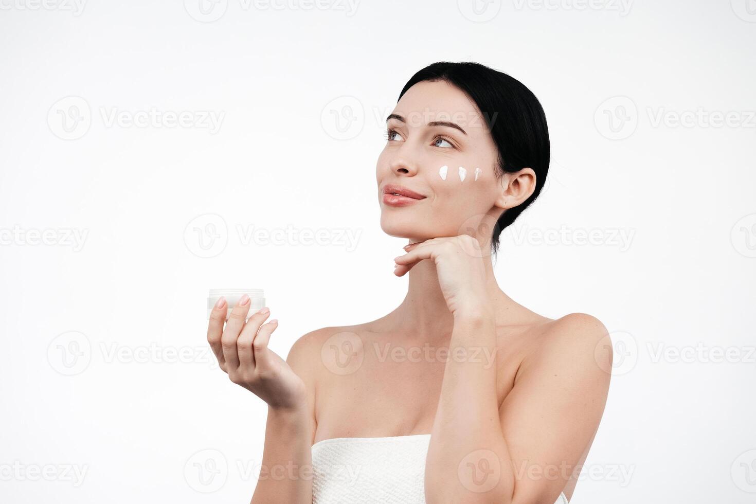 Beauty portrait of brunette woman in white towel, applies cream to the face. Skincare cleansing eco organic cosmetic spa relax concept. Free copy space for text, banner. Cosmetics advertising photo