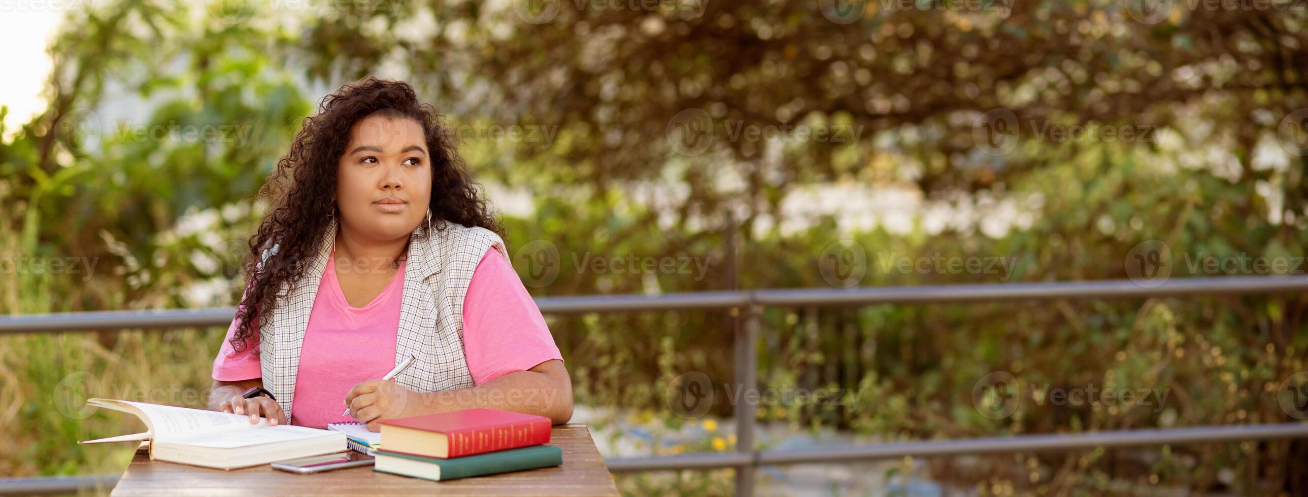 A young student sits at a table outside, surrounded by greenery, focused on her studies. She has books and notes open, enjoying the sunshine while working diligently on her assignments, panorama photo