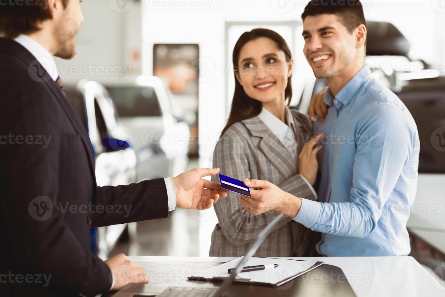 Buying Or Renting Car. Beautiful Couple Giving Credit Card To Vendor Standing In Dealership Office photo